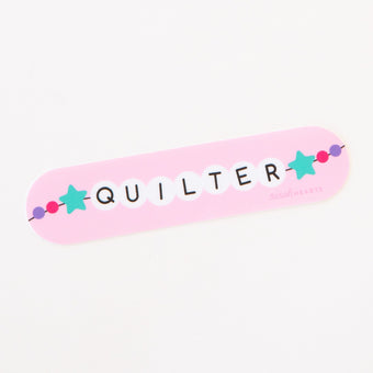 A light pink sticker featuring the word 'QUILTER' in a friendship bracelet beads and a teal star pattern.