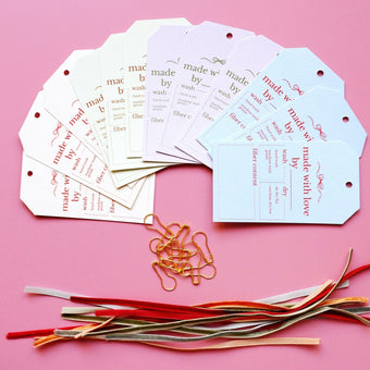 A set of handmade gift tags in cream, white, and light blue, printed with 'made by' and 'fiber' text, arranged on a pink background with gold paperclips and colorful thread spools.