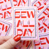 A hand with glittery red nail polish holds a die-cut sticker that reads 'SEW ALL DAY' in bold red letters on a pink patterned background. Multiple identical stickers are scattered in the background.
