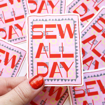 A hand with glittery red nail polish holds a die-cut sticker that reads 'SEW ALL DAY' in bold red letters on a pink patterned background. Multiple identical stickers are scattered in the background.